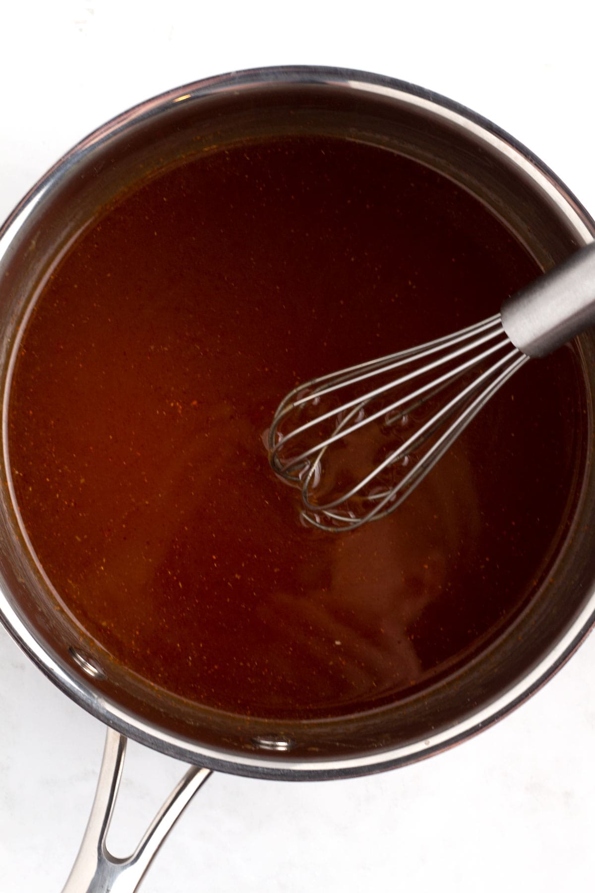 Overhead view of a silver pot with a brown glaze mixed together and a whisk resting on the edge of the pan.