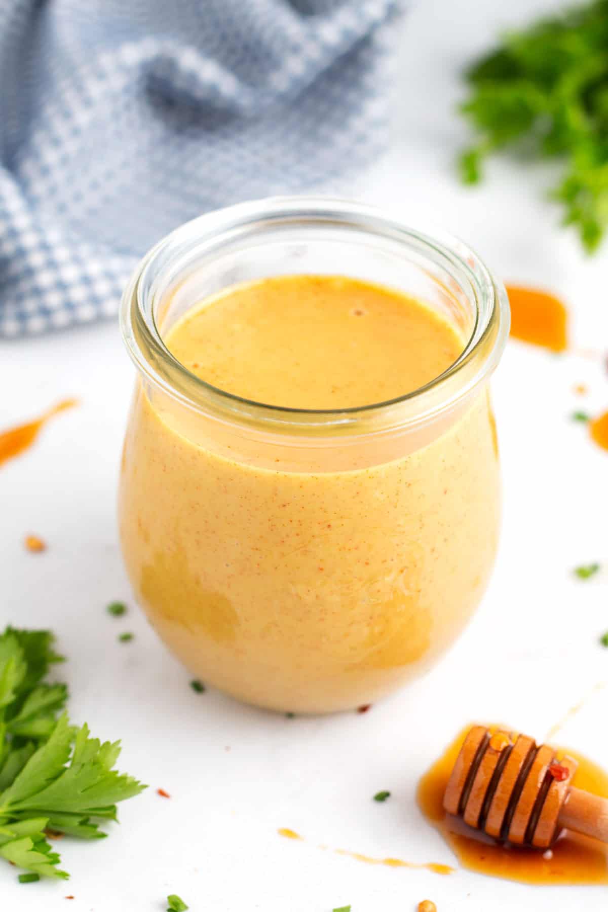 A glass jar of mustard sauce with parsley and a honey stick in front of the jar, a blue towel behind the jar, and drips of honey on the counter.