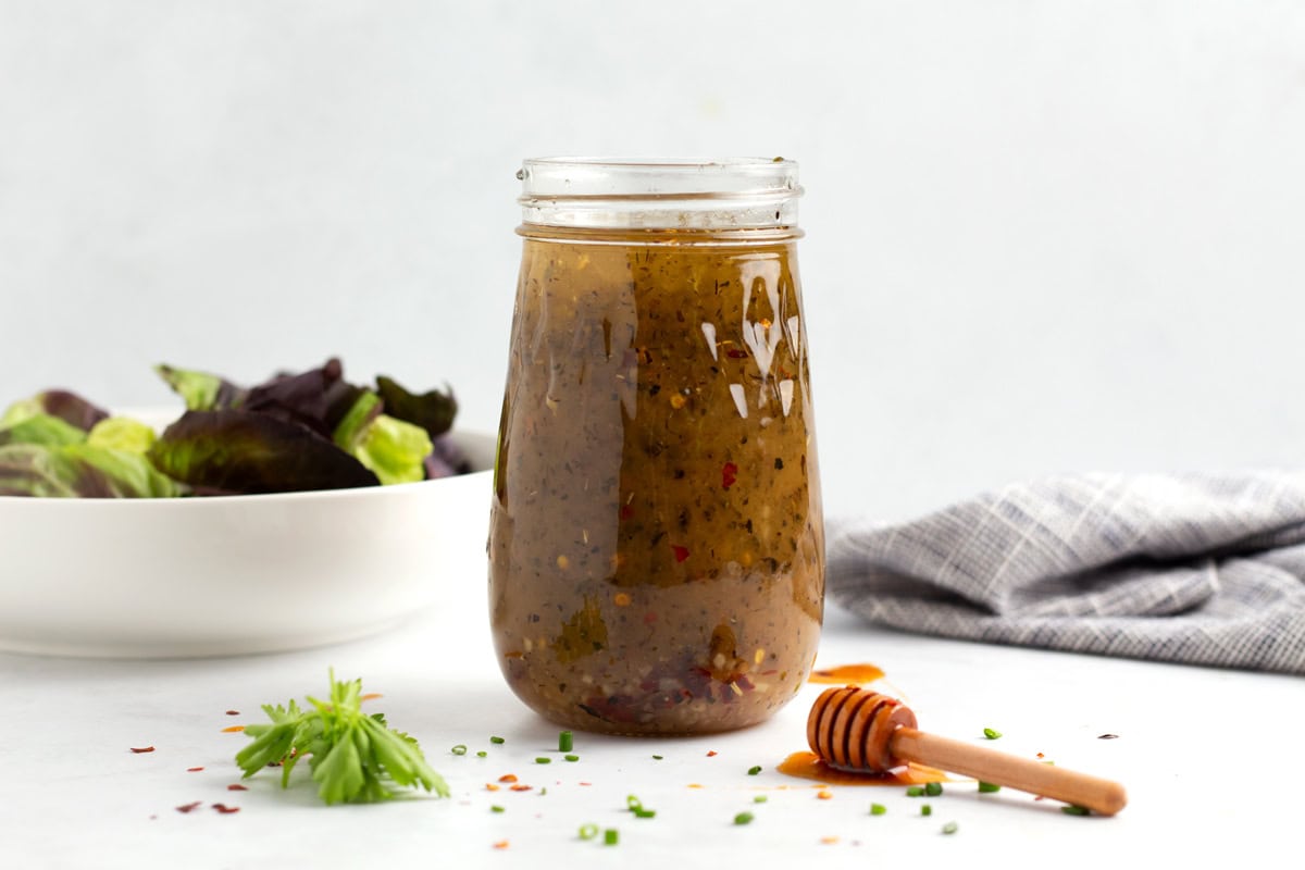 A full jar of spicy sweet vinaigrette with a bowl of lettuce, a blue towel, and a honey stick around the jar.