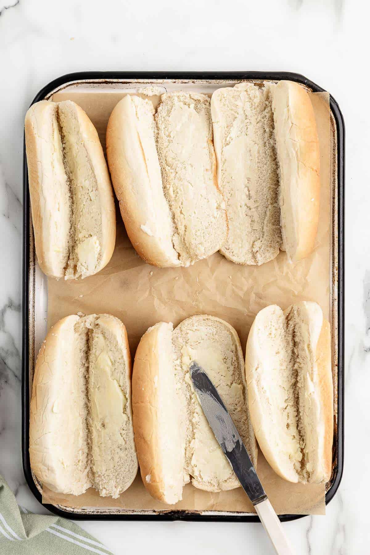 Overhead view of 6 hoagie rolls spread with softened butter on a cookie sheet with a butter knife laying on one roll.