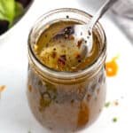 Jar of hot honey dressing with a spoon being lifted out of it and a bowl of green lettuce in the top left corner.