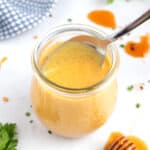 A square image of a jar of hot honey mustard with a spoon coming out of it, parsley in front of the jar, a blue towel behind the jar, and drips of honey on the counter.
