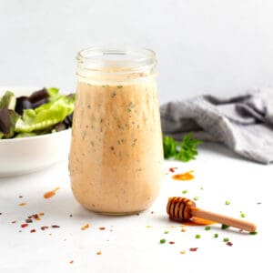 Salad dressing jar full of hot honey ranch with bowl of salad behind and chili flakes throughout the scene.