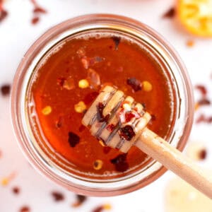 Overhead view of a honey stick dripping with honey and sprinkled with red pepper flakes over a glass jar.