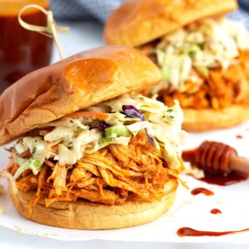 Two shredded chicken sandwiches on a piece of parchment paper with red sauce drips and a jar of honey sauce in the background.