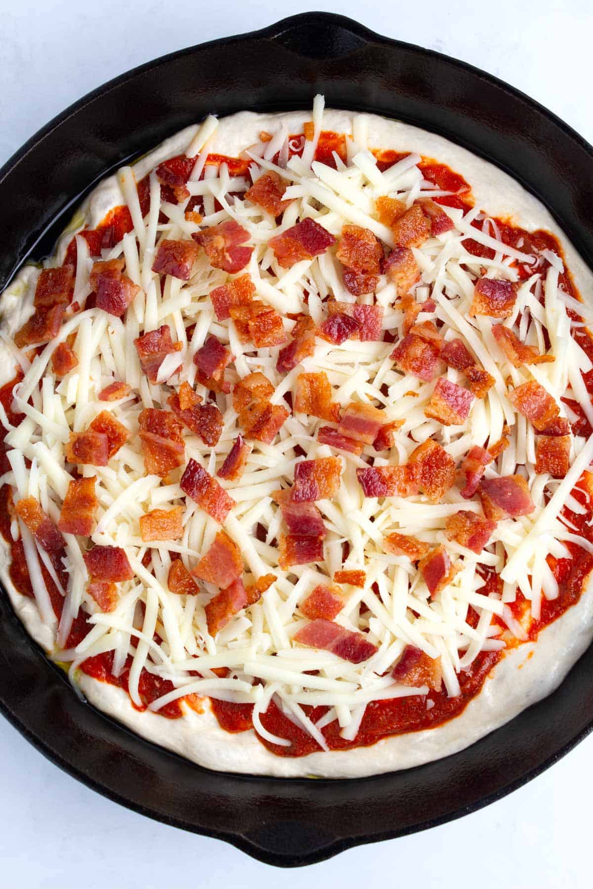 Dough spread out in cast iron pan topped with sauce, cheese, and bacon bits.