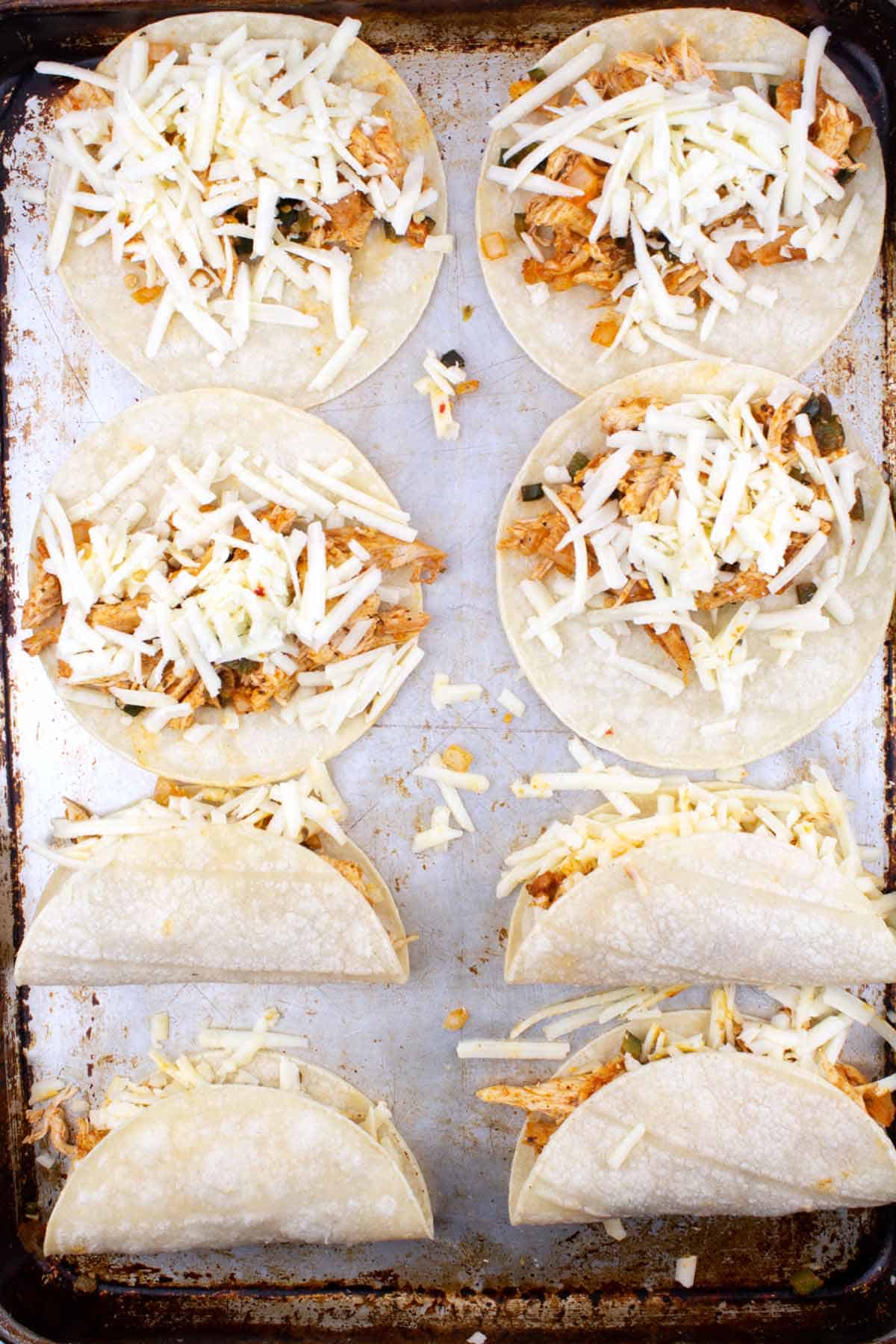 Eight tortillas topped with shredded meat and cheese on a cookie sheet; four of them are folded over in half.