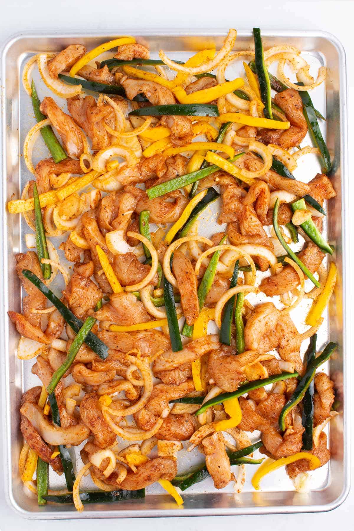 Raw sliced chicken, peppers, and onions tossed with seasonings on a large silver baking tray.