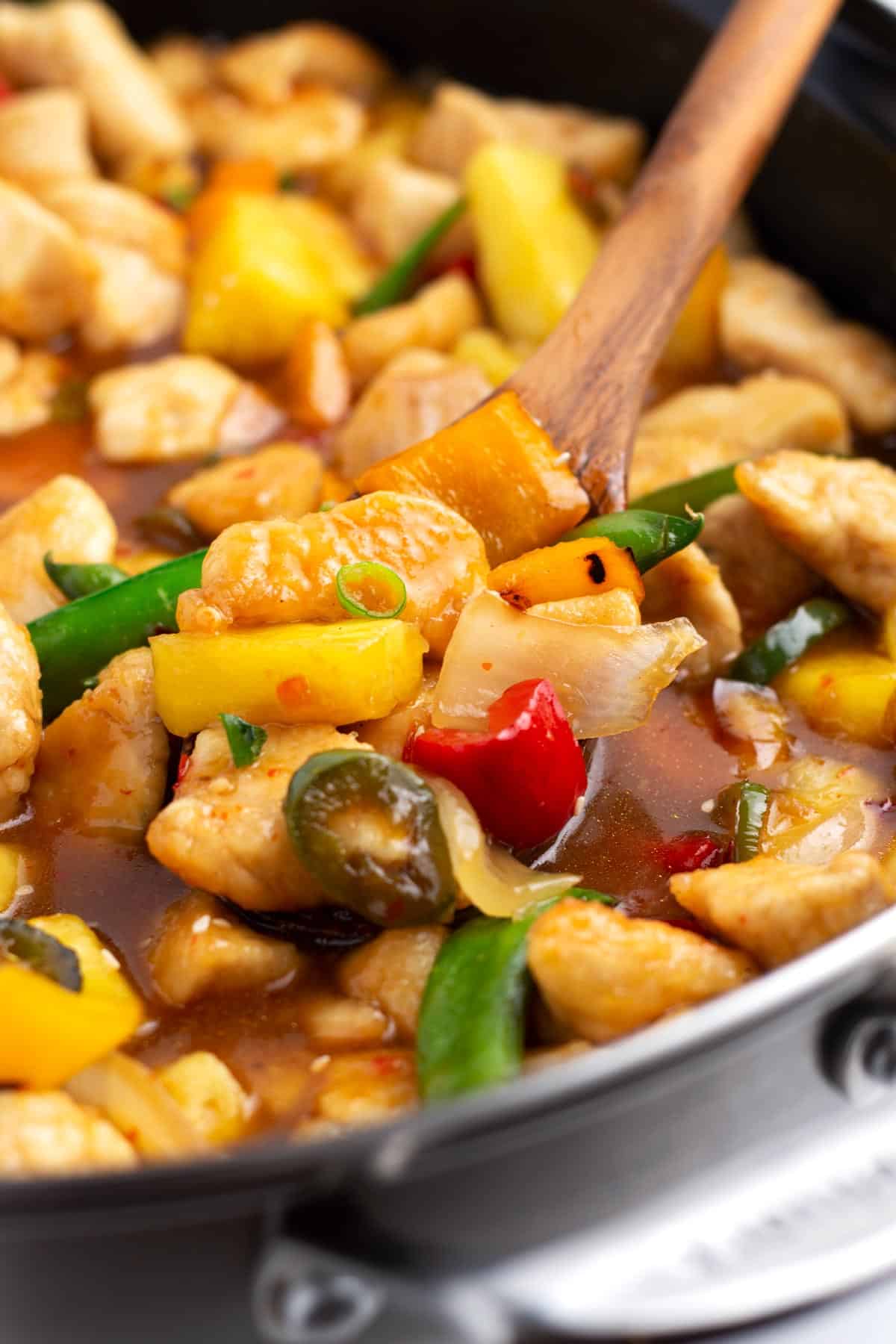 A black skillet of sweet and spicy stir fry with a wooden spoon resting on the right side.