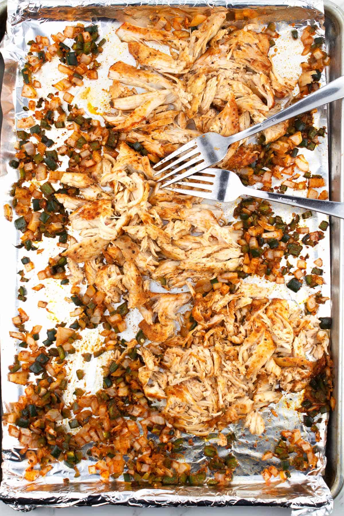 Two forks shredding chicken and vegetables on a baking tray lined with foil.