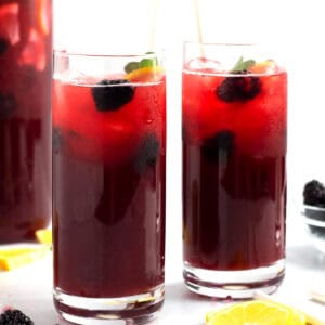 Two clear glasses full of a purple liquid with straws, blackberries, and slices of lemon around the jars.