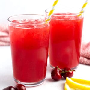 Two glass cups of a summer drink with yellow straws, fresh cherries, and lemon wedges around the scene.