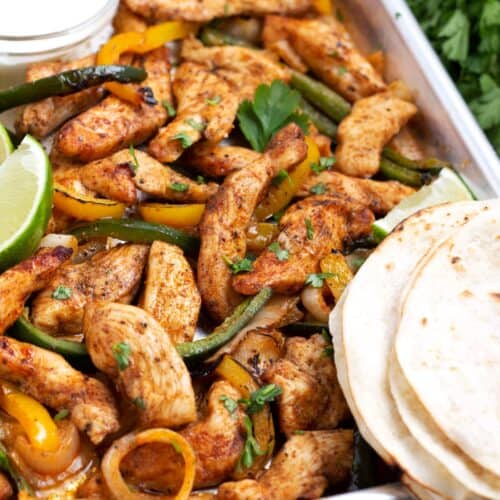 Seasoned chicken strips and roasted bell peppers with flour tortillas in the foreground and lime wedges in the background.