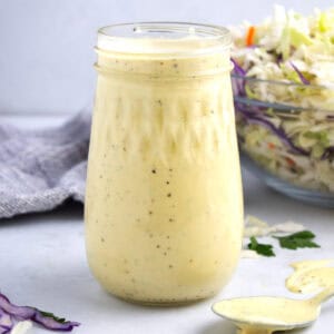 Fluted jar with creamy homemade coleslaw dressing and spoon laying on counter.