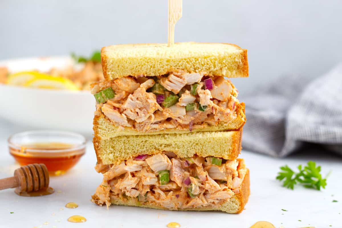 Two pieces of bread filled with sweet and spicy chicken salad on countertop with serving bowl, napkin, and spicy honey.