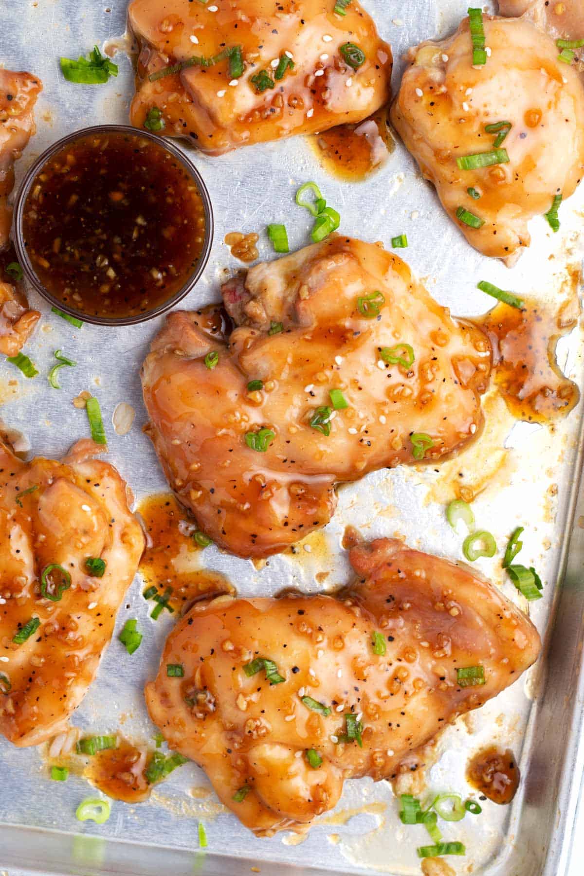 Overhead view of a baking sheet full of baked chicken thighs covered in a brown sauce with sliced green onions and extra sauce scattered around the scene.