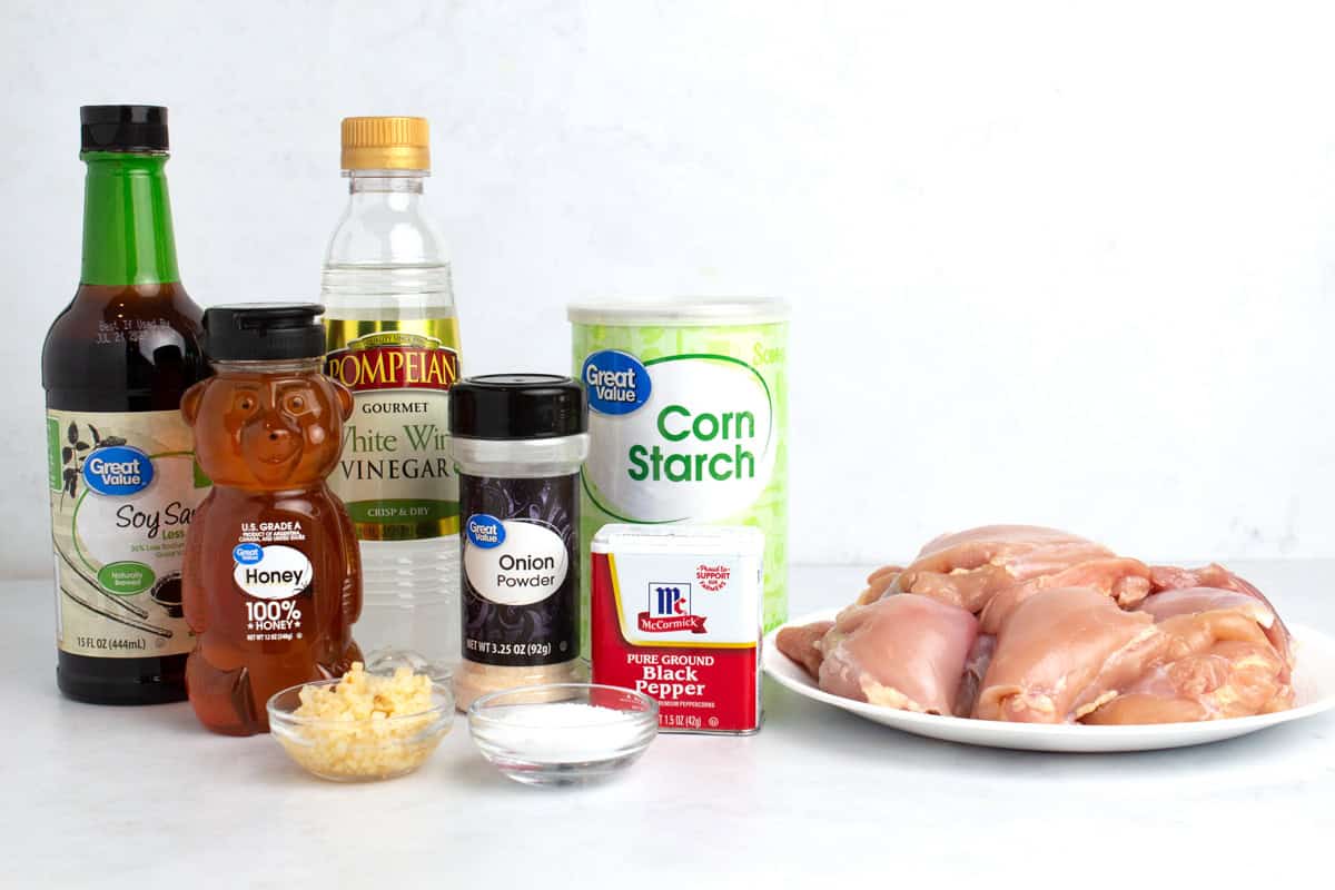 Raw chicken, soy sauce, honey, white vinegar, garlic, onion powder, cornstarch, salt, and pepper on a white background.