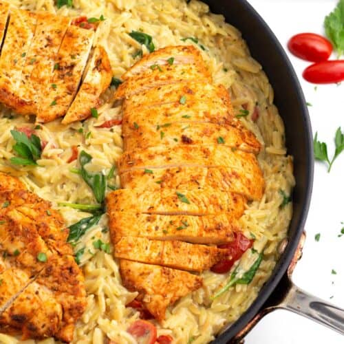 A square image of a skillet with creamy orzo, sliced chicken, tomatoes, and spinach.