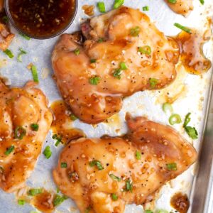 An overhead view of sauce covered chicken on a silver sheet pan sprinkled with green onions and sesame seeds.