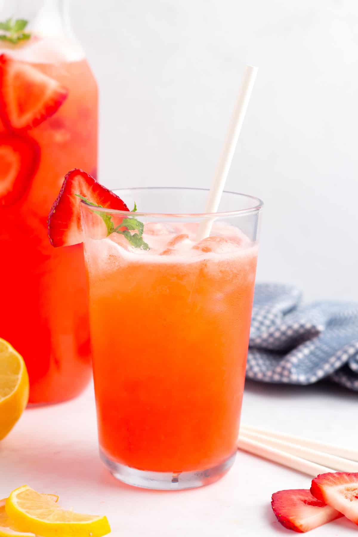 Tall cold glass of fresh lemonade with strawberries with lemons, strawberries, and straws on the countertop.