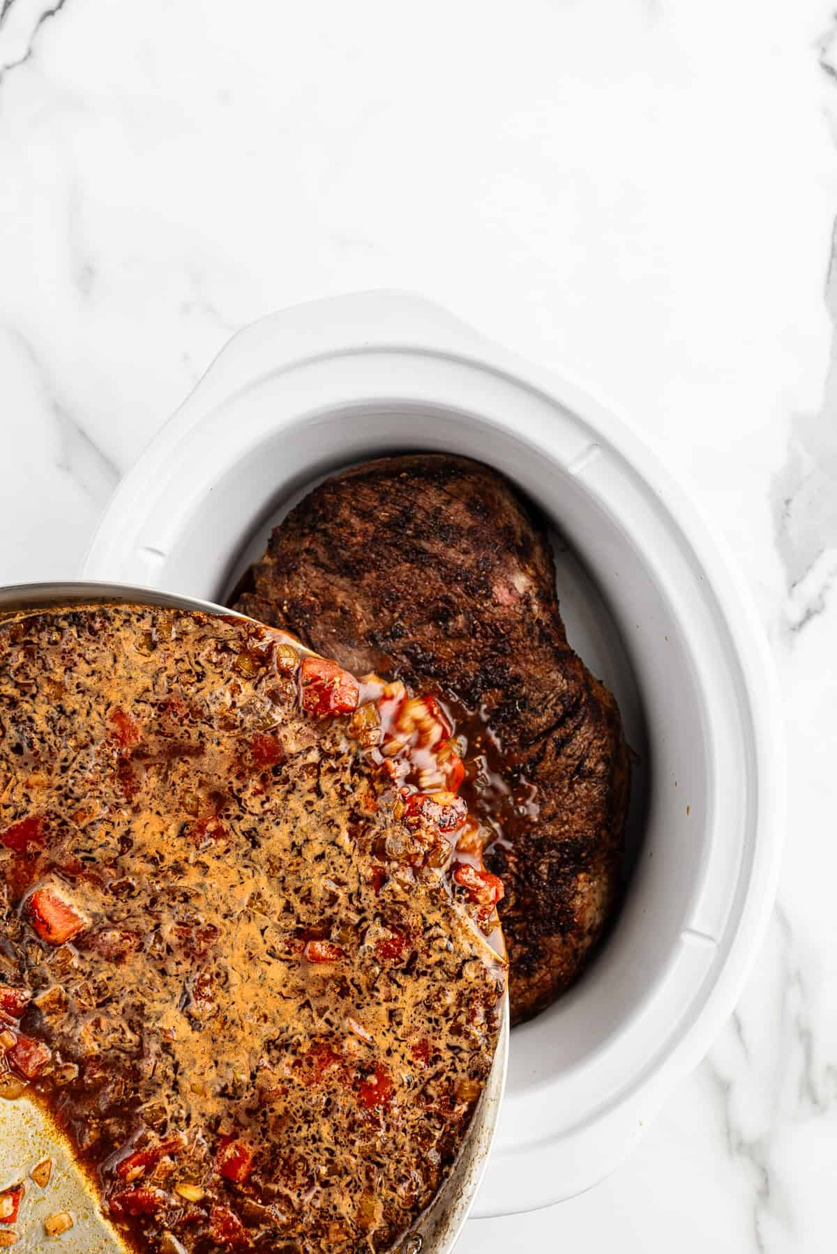 Cooked liquids being poured from a skillet into a white crock pot on top of a roast.