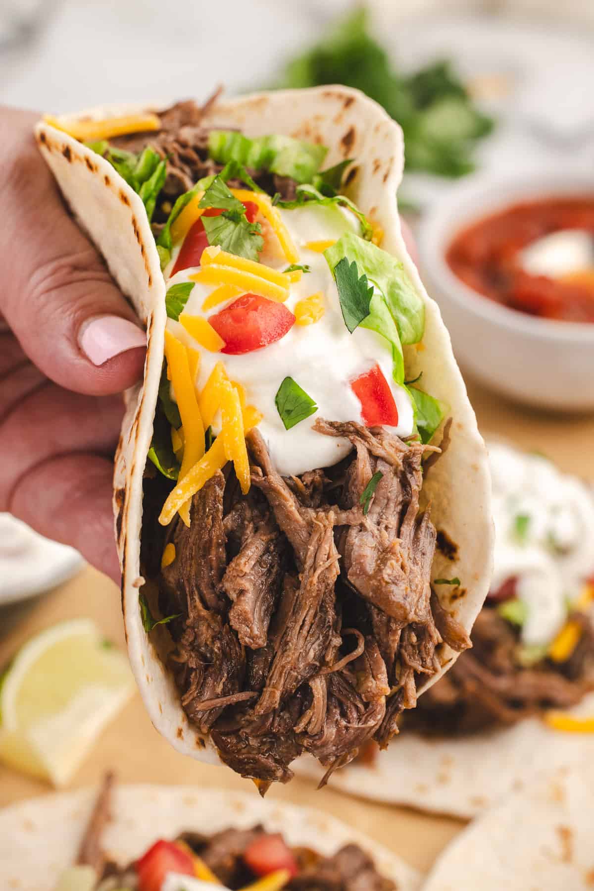 A hand holding a very full taco full of shredded beef and topped with sour cream, cheese, and veggies.