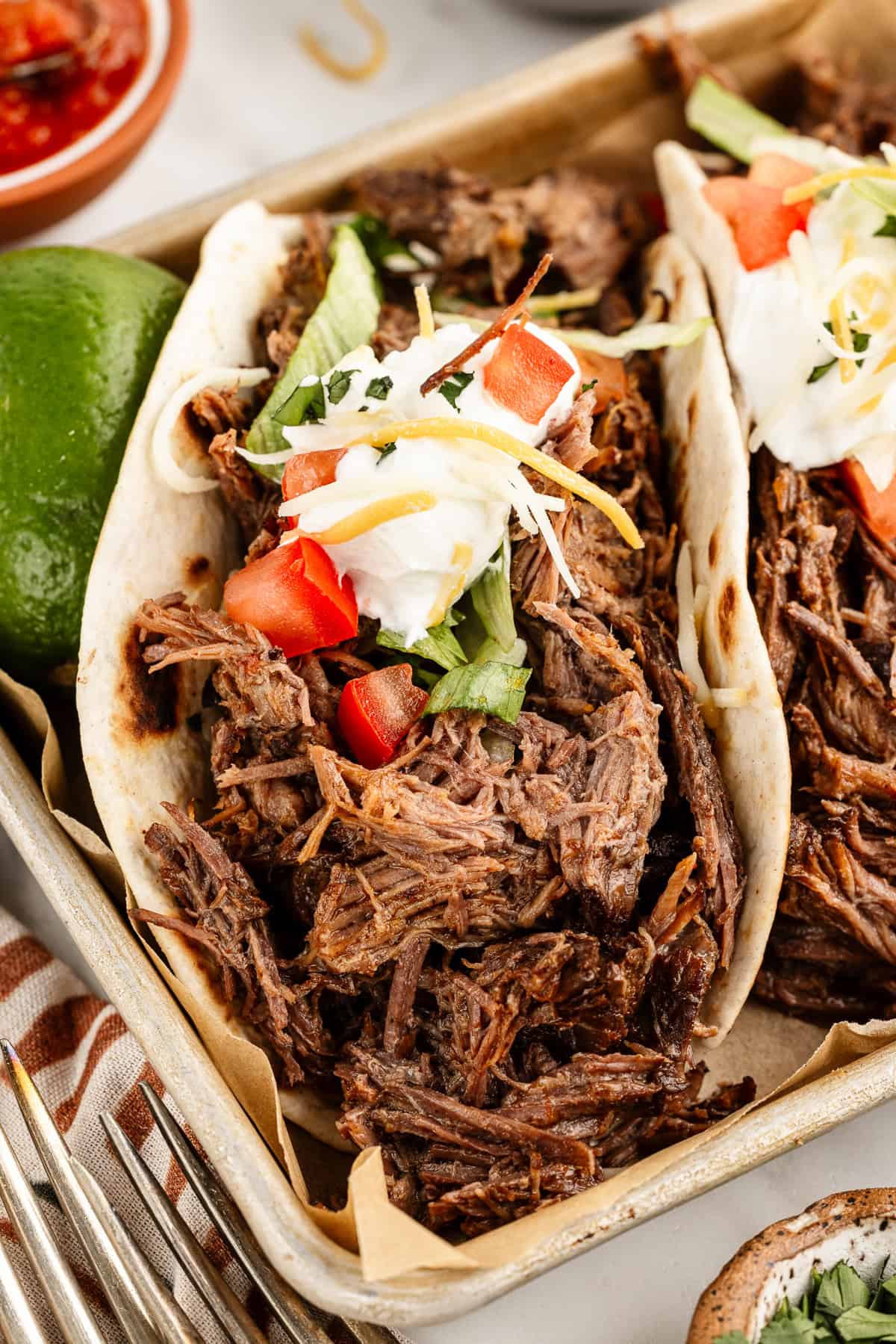 A close up image of a shredded beef taco with toppings sitting on top of a cookie sheet lined with brown parchment paper.