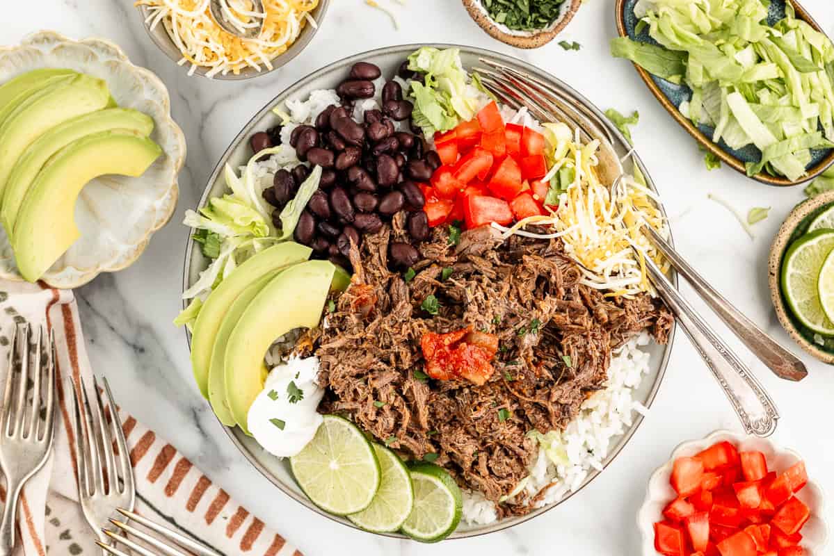 A plate full of rice, beans, meat, cheese, limes, tomatoes, and avocado, surrounded by more toppings and forks on a marble countertop.