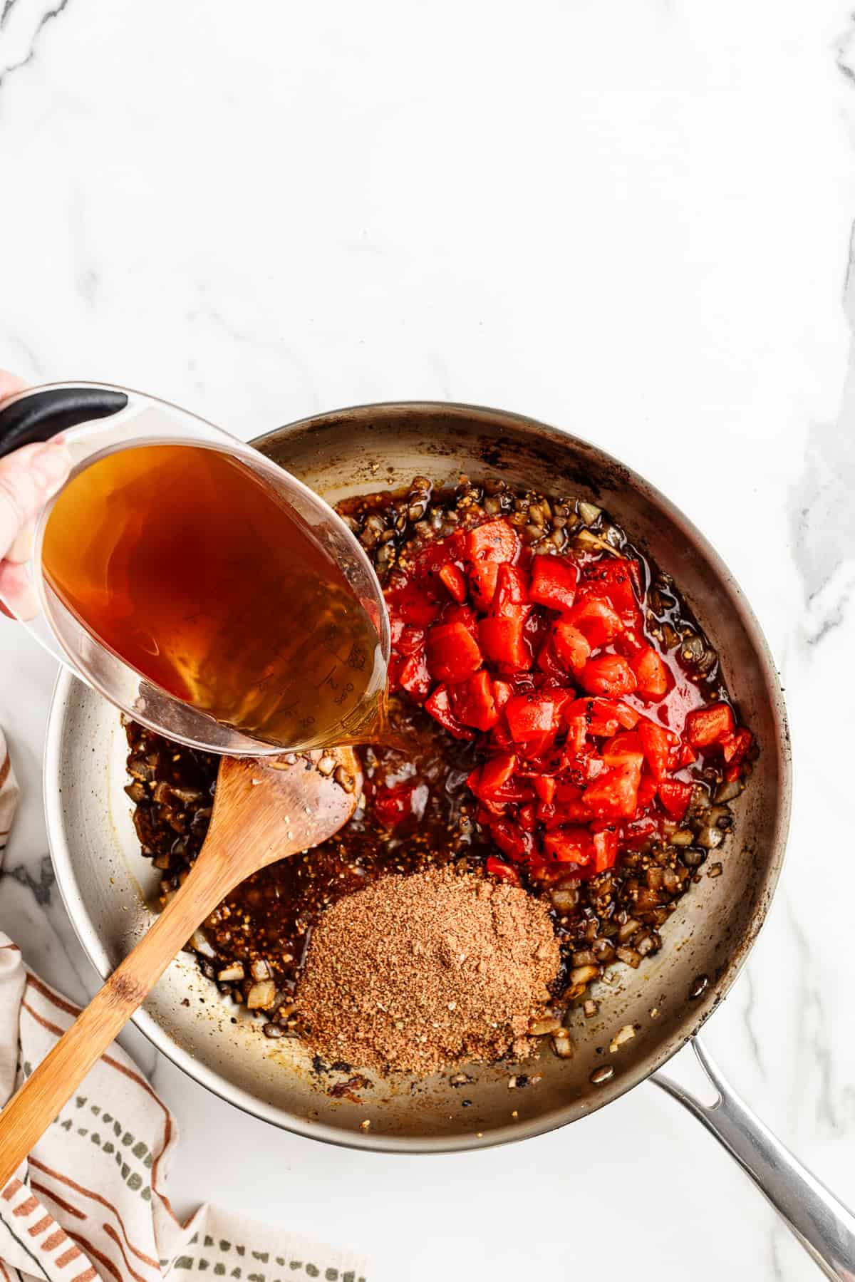 A hand pouring broth into a silver skillet that has a wooden spoon, tomatoes, onions, and seasoning inside.