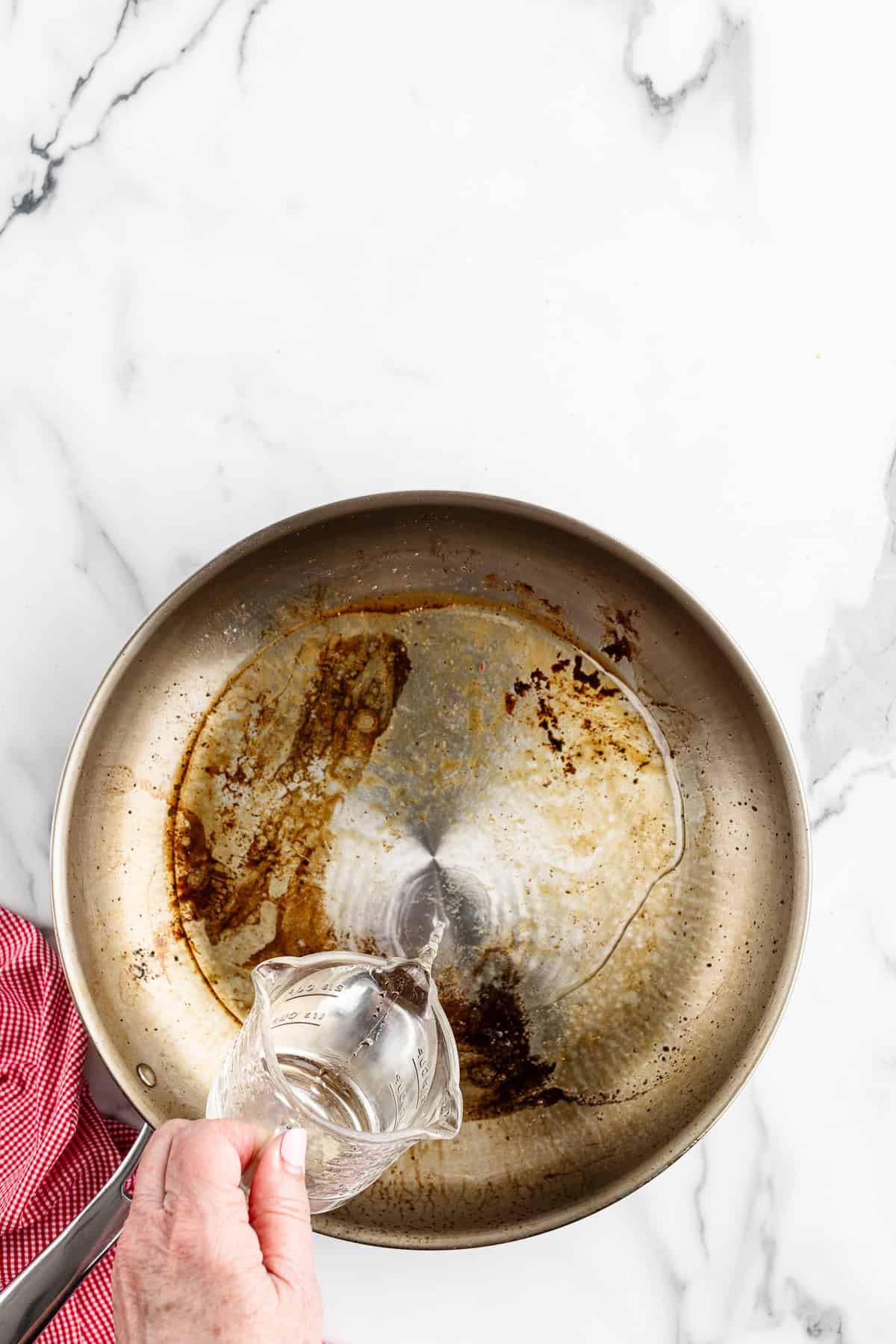 A hand pouring a glass of water into a stainless steel skillet that has brown searing marks on it.