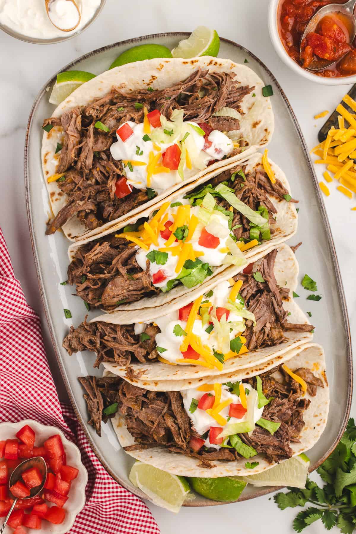 Overhead of an oval platter full of tacos with a bowl of tomatoes on the left, a bowl of salsa and cheese on the right, and a dish of sour cream on the top.