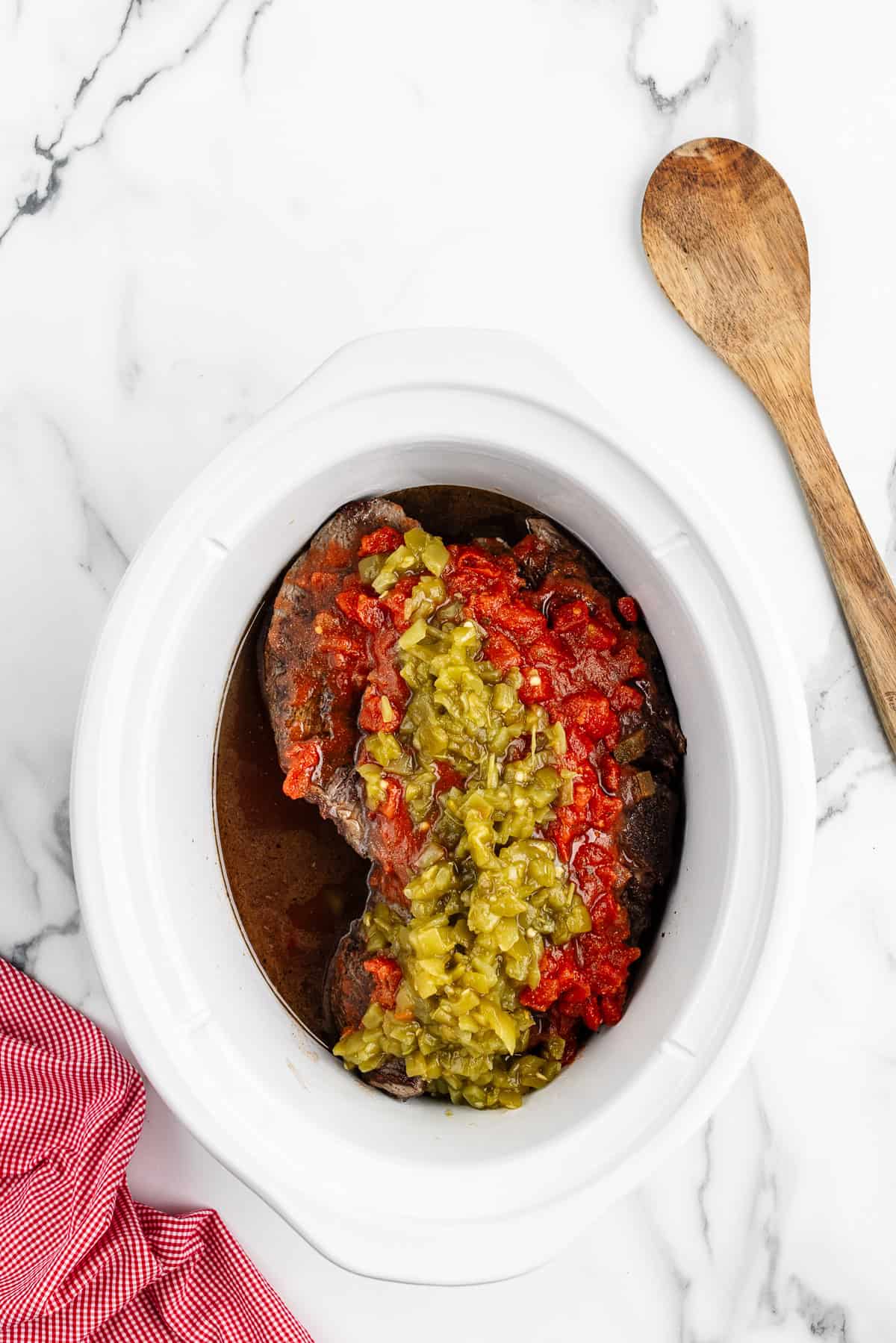 A white crock pot filled with meat, broth, chunky salsa, and green chilis. There is a red checkered towel and wooden spoon on either side of the pot.