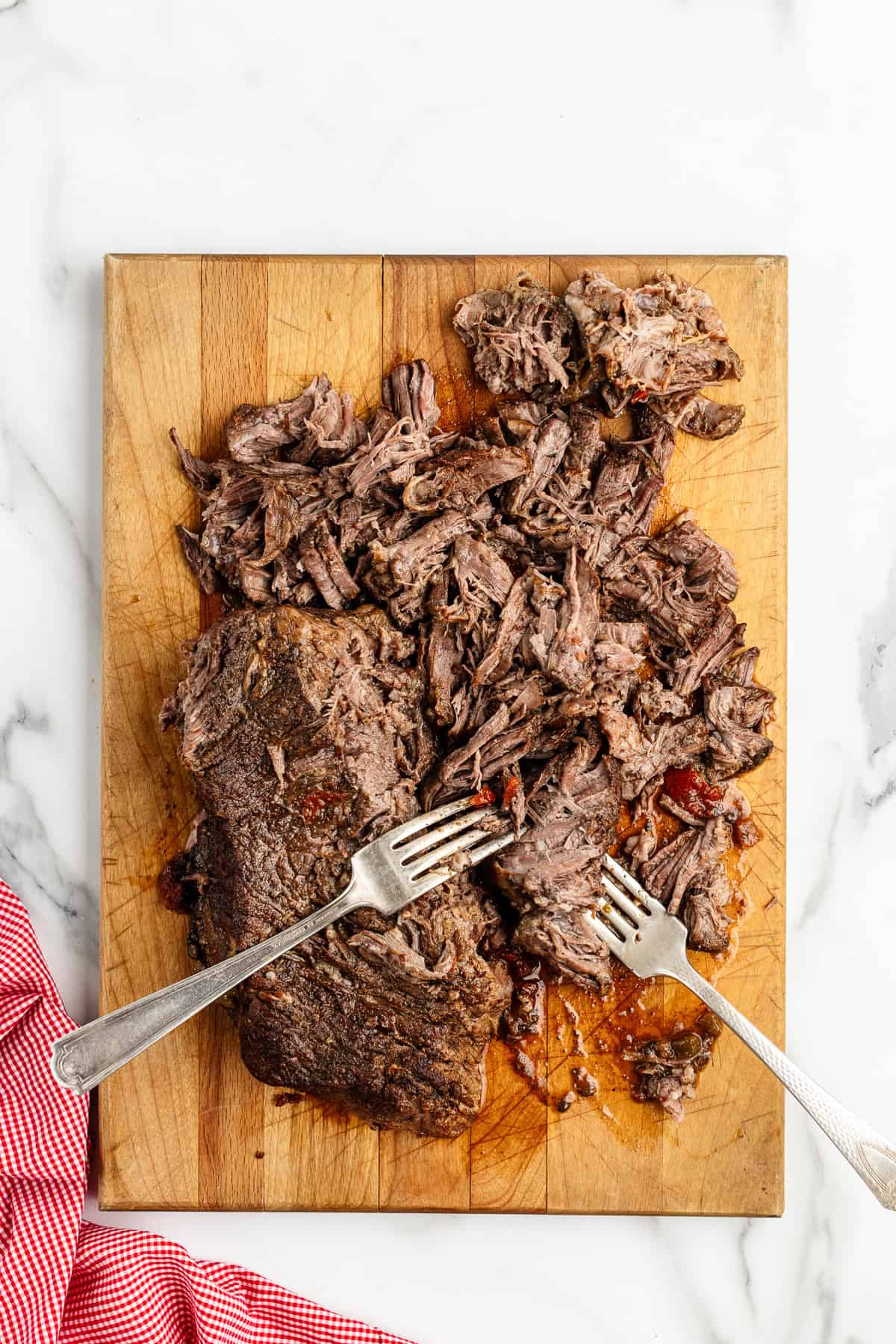 A wooden cutting board with a chunk of cooked roast being shredded with two forks.