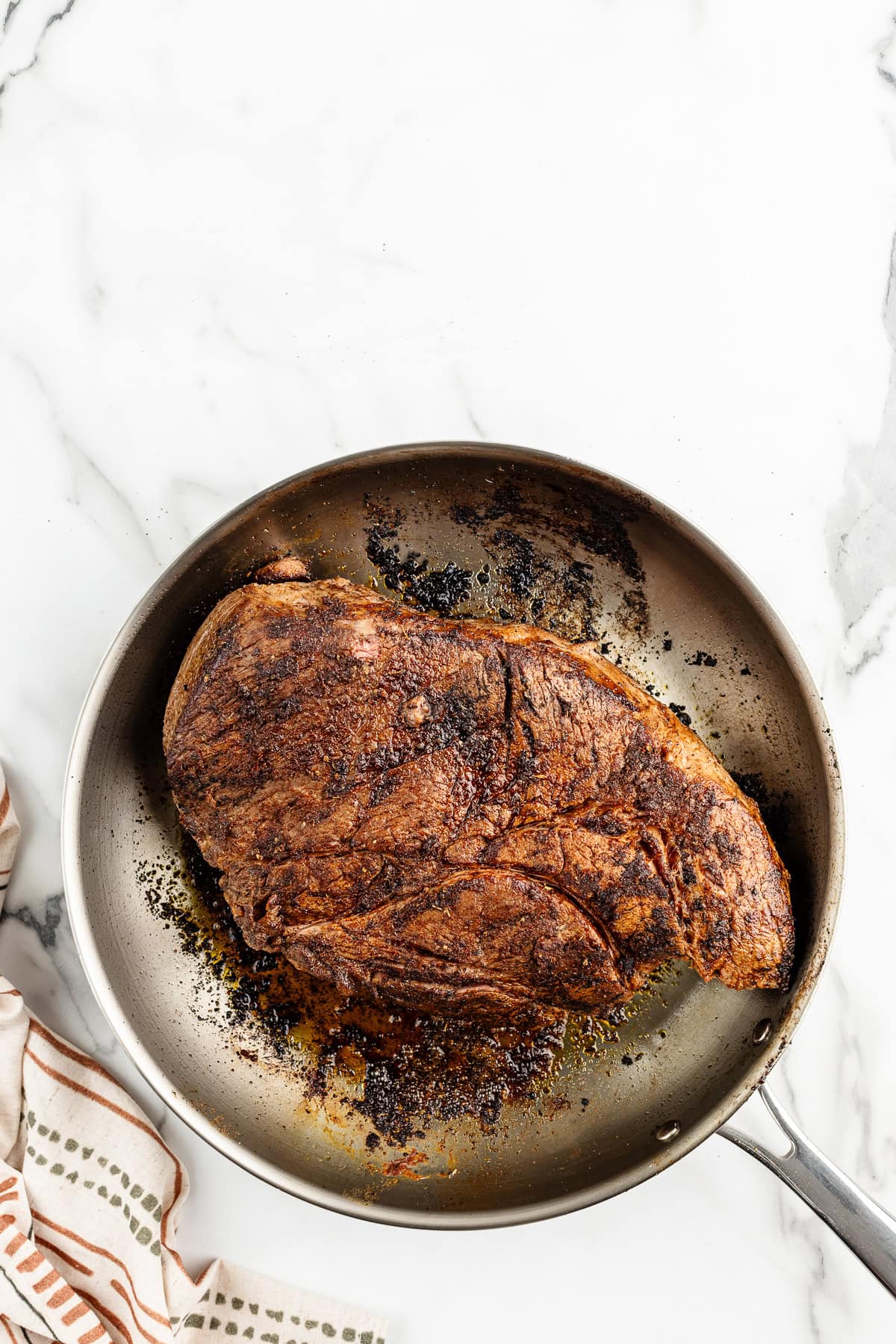 A golden brown chuck roast searing in a silver skillet with a towel in the bottom left corner.