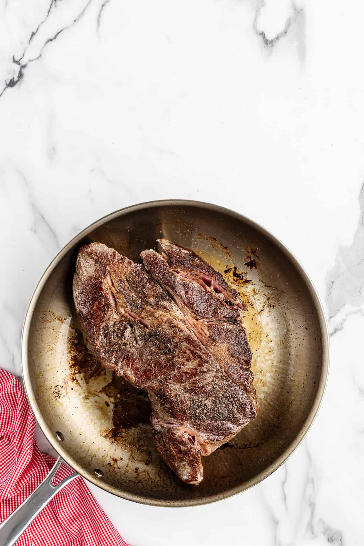 A large chunk of seared beef in a stainless steel skillet with a red checkered towel on the side.