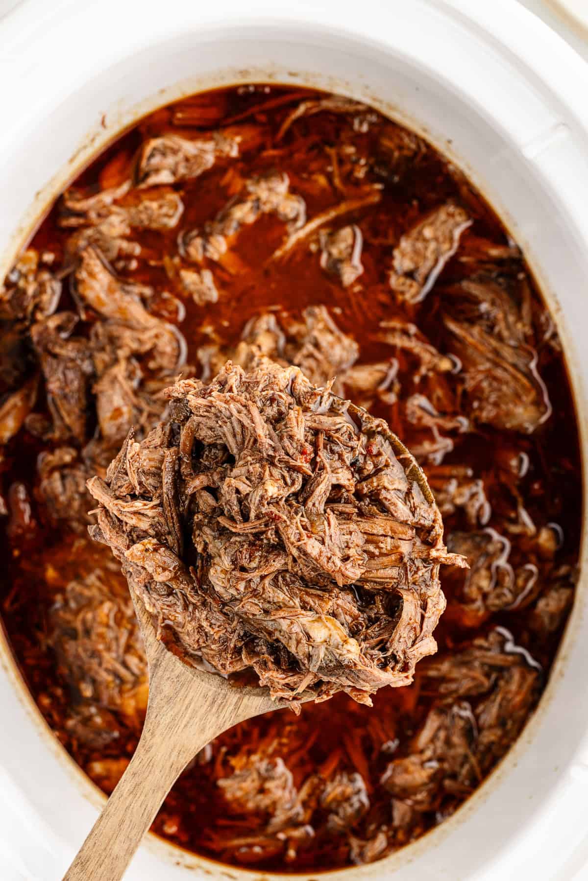 A spoon holding a scoopful of cooked meat over the top of a white crock pot.