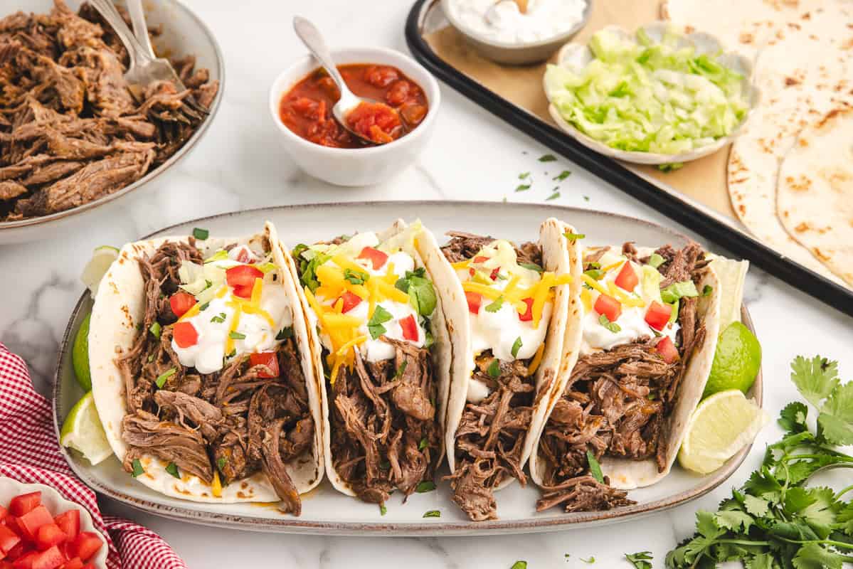 A horizontal image of an oval platter lined with four beef tacos surrounded by a bowl of shredded meat, a bowl of salsa, extra tortillas, and pieces of cilantro.