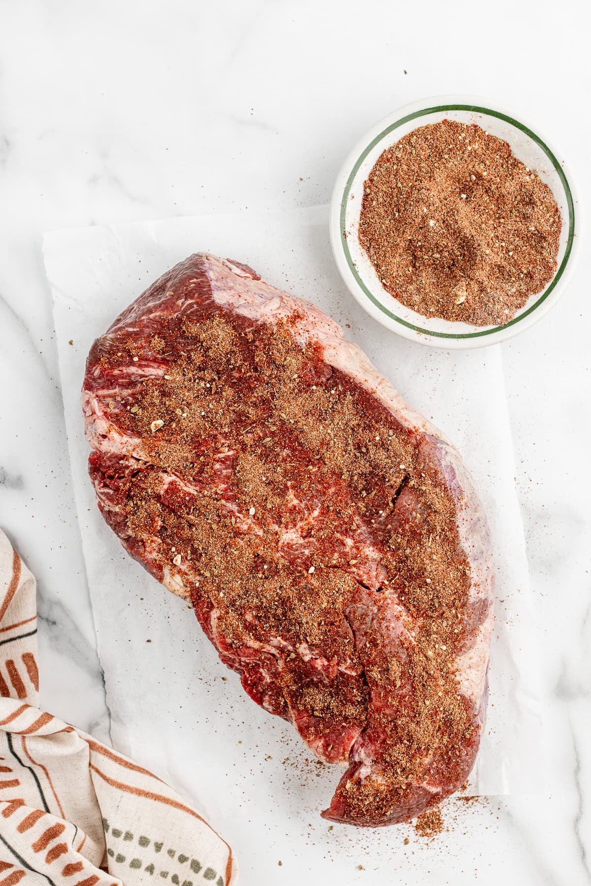 A raw roast on a white counter top sprinkled generously with a reddish brown seasoning mixture.