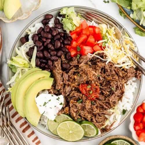 A tan plate full of rice, beans, beef, and Mexican toppings with forks and extra toppings on on the sides.