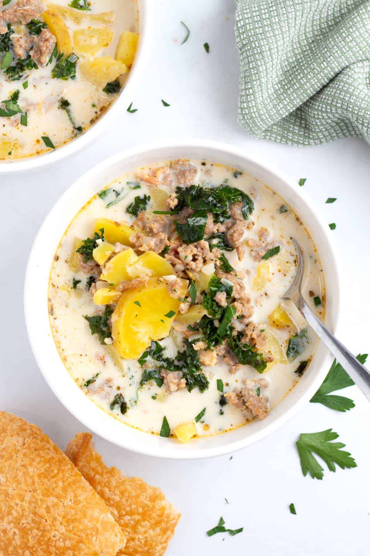 Two bowls of sausage and potato soup with kale on tabletop with crusty bread and napkin.