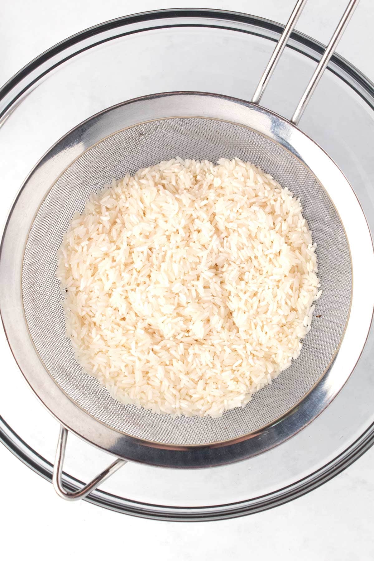 White long-grain rice in a fine mesh strainer over a bowl rinsing off the starch.