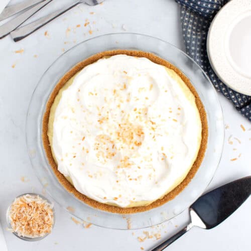 A coconut dessert topped with whipped cream and a serving utensil, forks, and plates on the side.