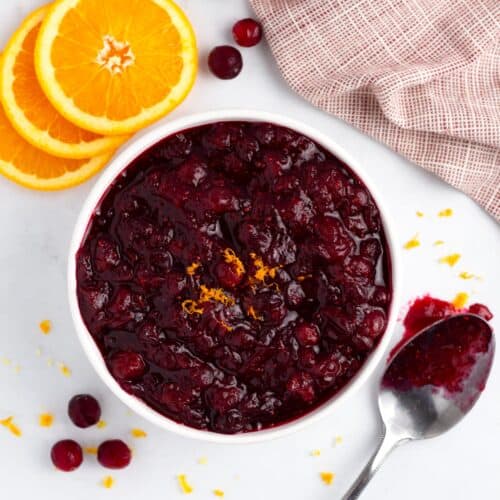 An overhead view of a white bowl of cranberries with orange slices, a red towel, a spoon all around it.