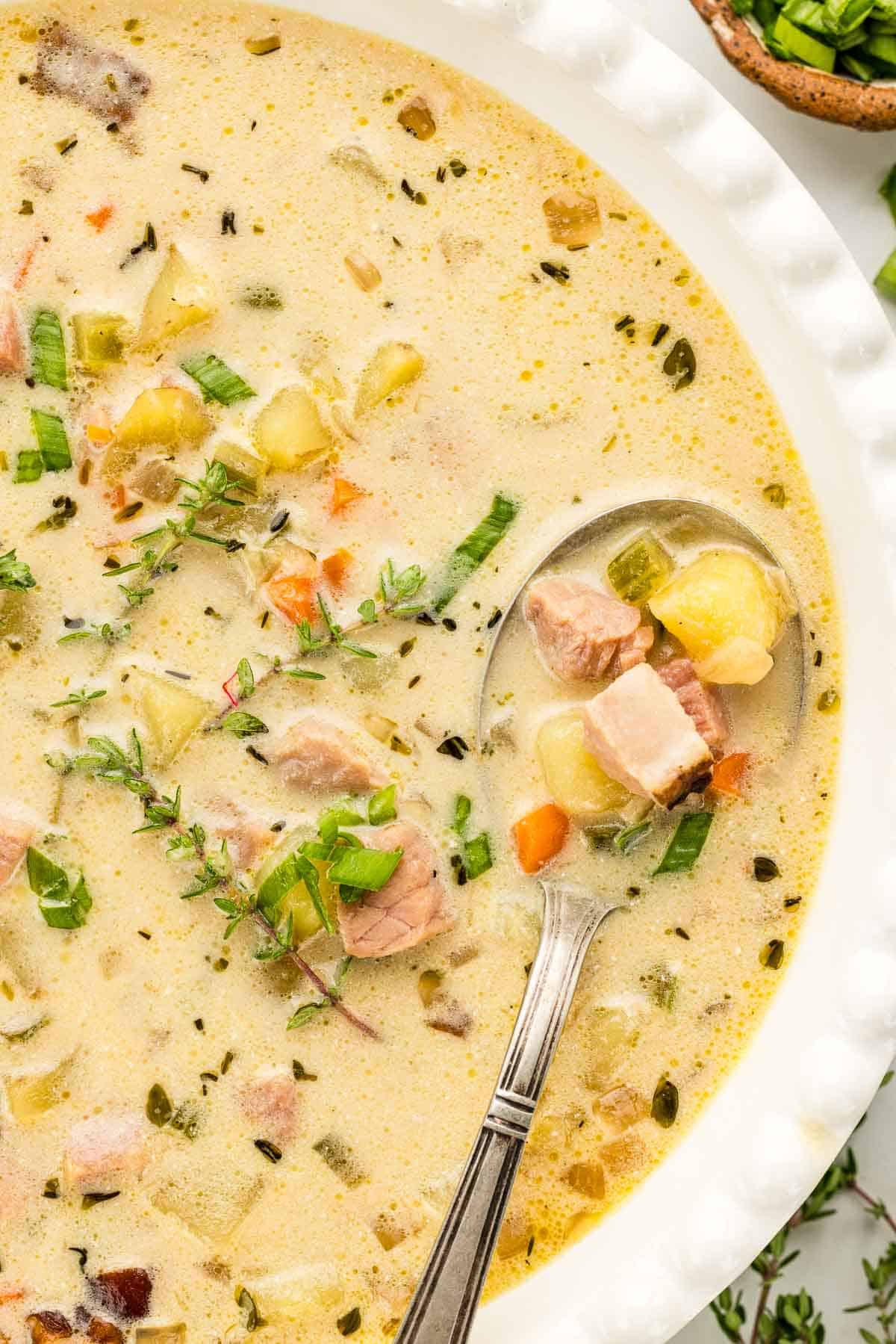 Bowl with spoon inside of creamy broth filled with potatoes, vegetables, and ham.