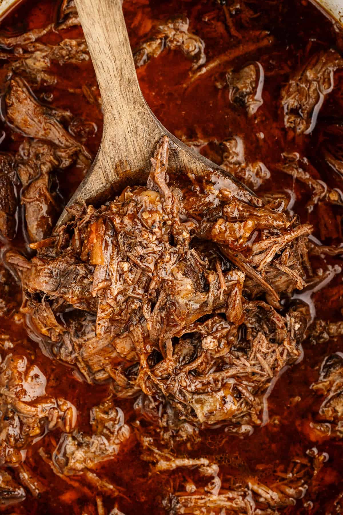 A close up view of a wooden spoon submerged in pulled beef that is coated in a dark red, liquidy sauce.