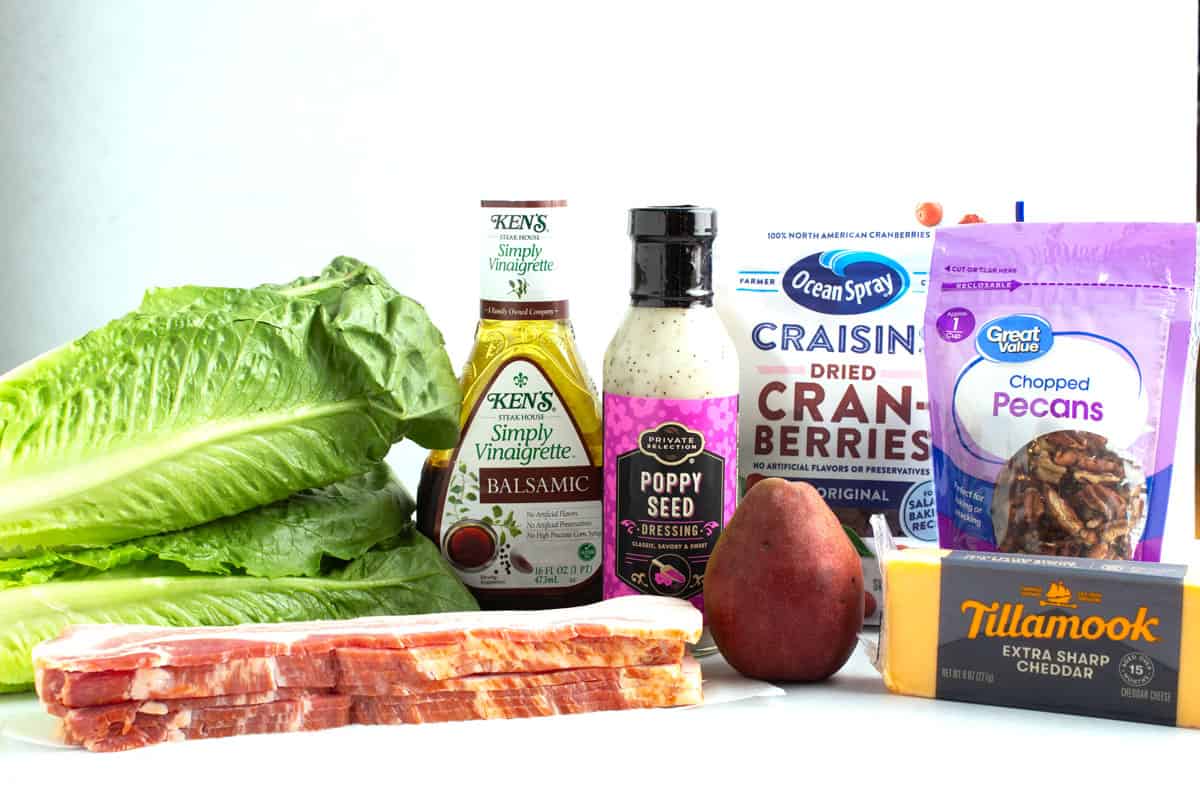 Ingredients for autumn chopped salad including romaine lettuce, bacon, red pear, sharp cheddar cheese, chopped pecans, dried cranberries, poppy seed dressing and balsamic dressing on the counter.