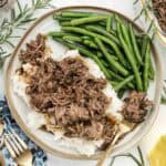 Shredded meat served over mashed potatoes with green beans on the side all plated on a white and gold dish with sprigs of rosemary and utensils on the sides.