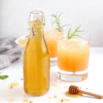 Two sweet and spicy cocktails and a glass container of hot honey simple syrup with honey drips on countertop.