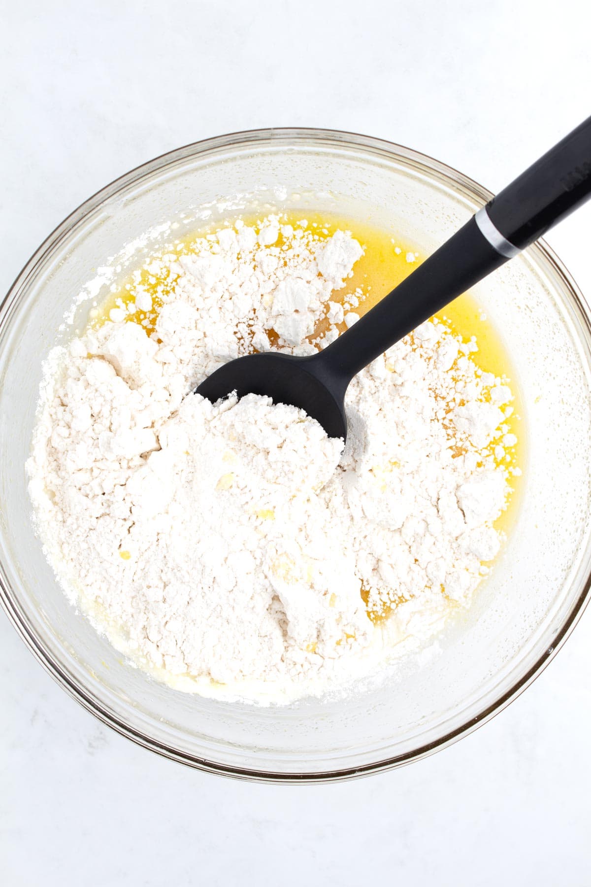 A glass bowl of batter with unstirred flour and a black spatula sitting in the bowl.
