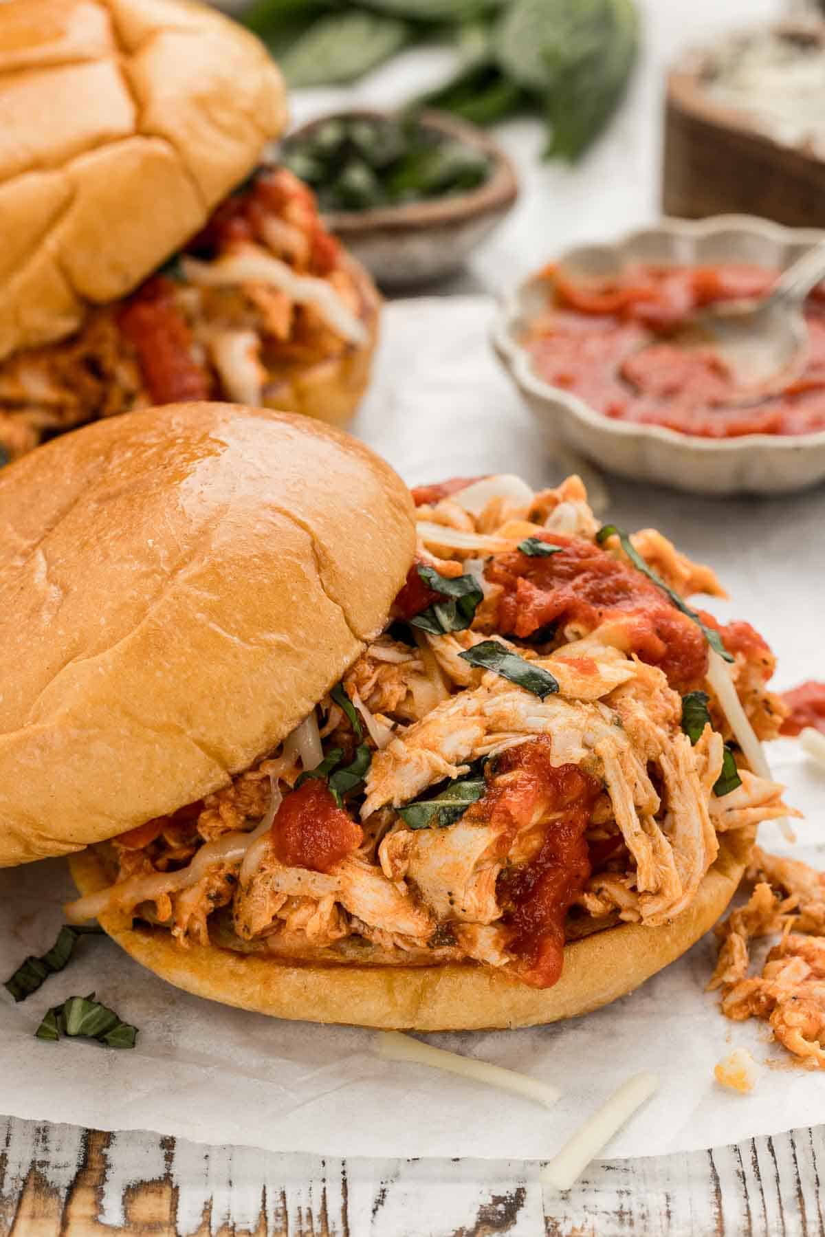A shredded chicken parmesan sandwich with the top bun sliding off with another sandwich next to it and a bowl of sauce and a bowl of herbs behind it.